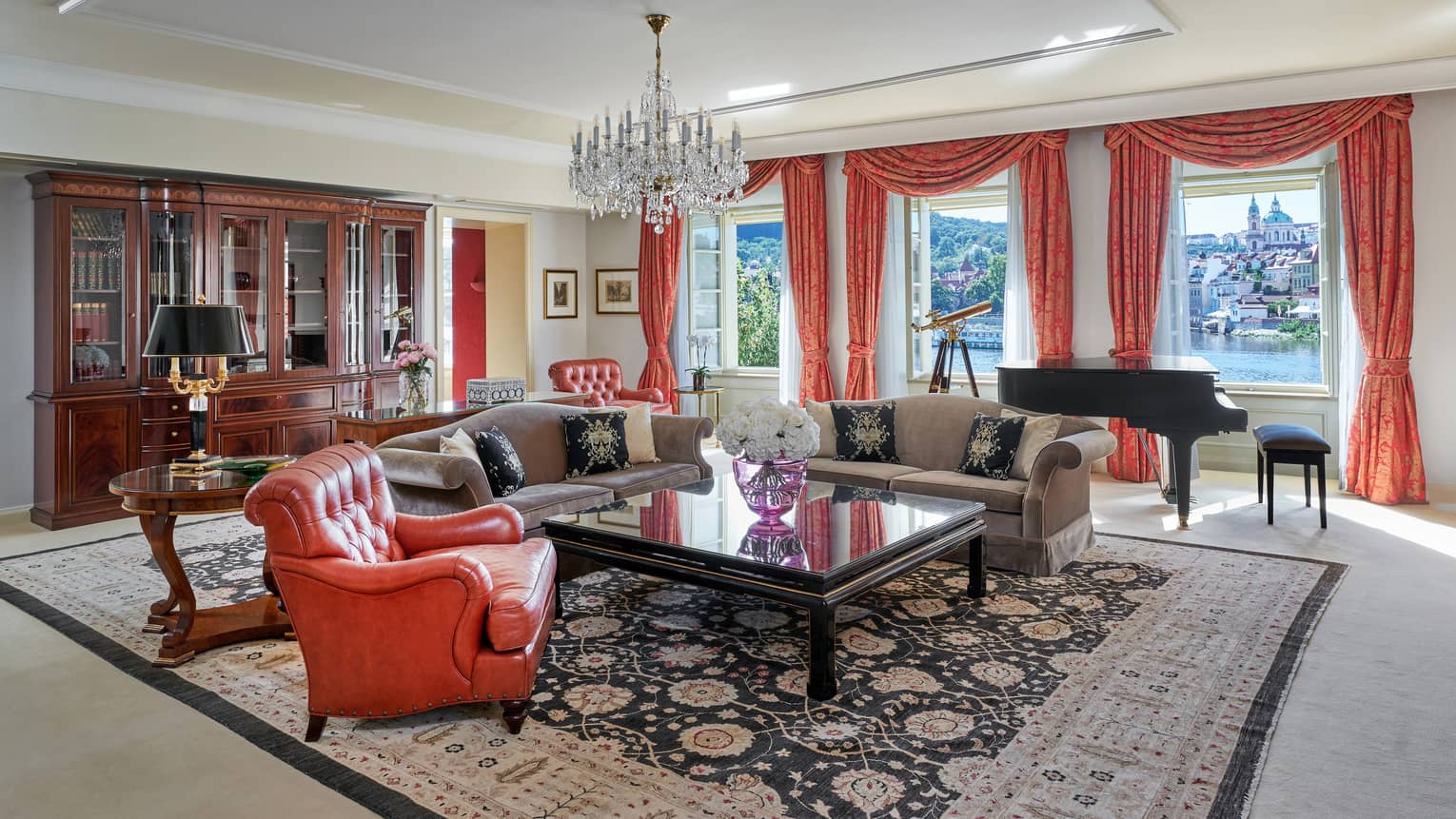 A luxury hotel suite's living room, with classic design, featuring a chandelier, red tapestry and a large square coffee table surrounded by two sofas and a red leather arm chair