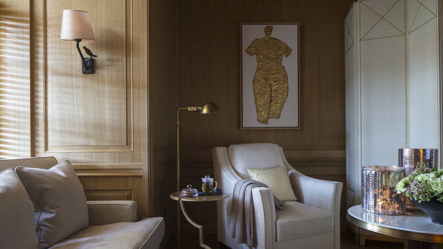 Sunlight streams through window to corner of sofa, white armchair with blanket on arm, reading lamp, art piece on wall