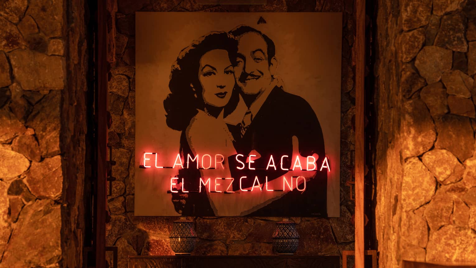 A neon sign with Spanish writing in front of a painting of two people hugging on a stone wall.