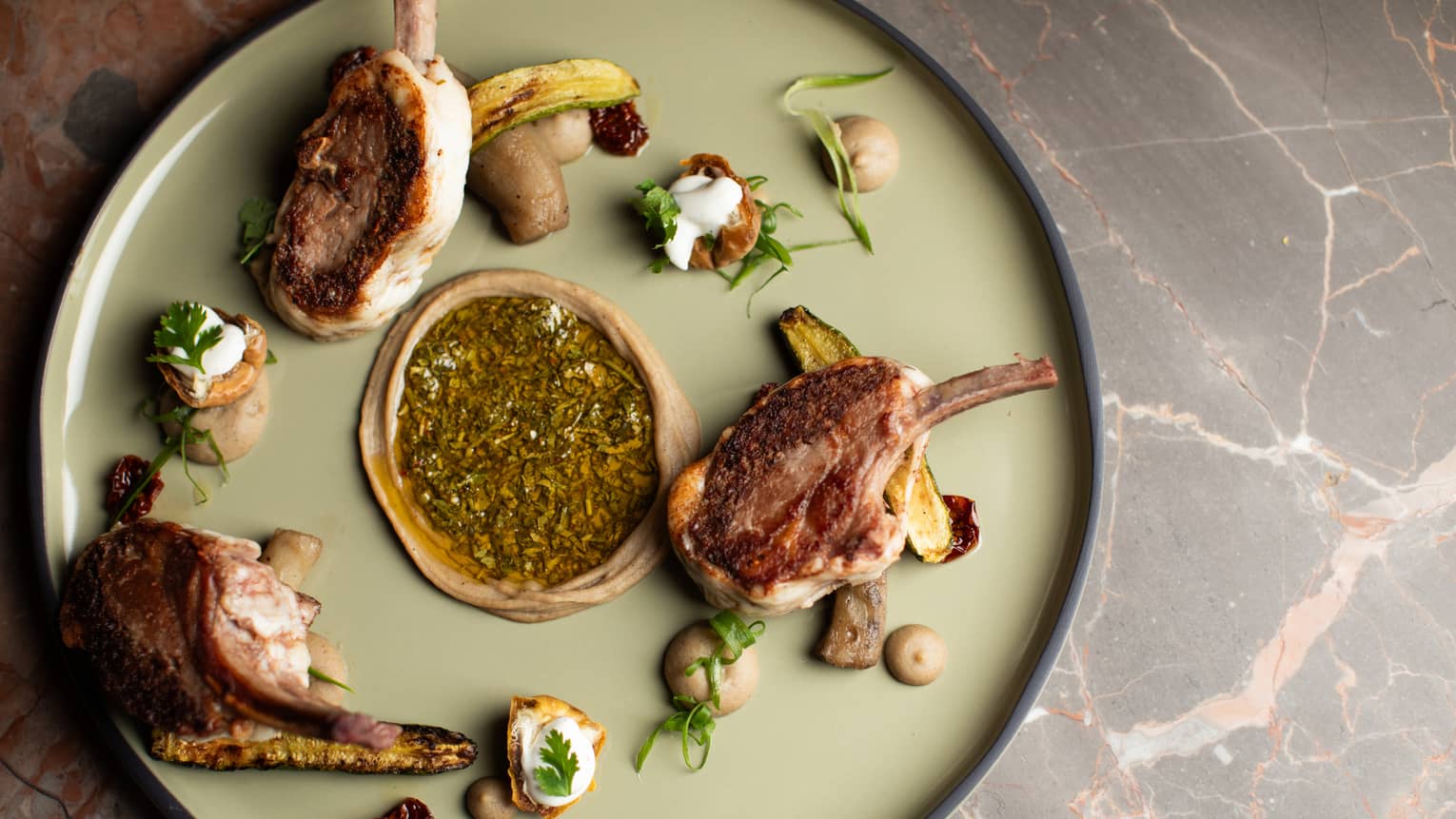 Three grilled lamb chops plated with eggplant and an assorment of garnishes on a large, round, light-green plate
