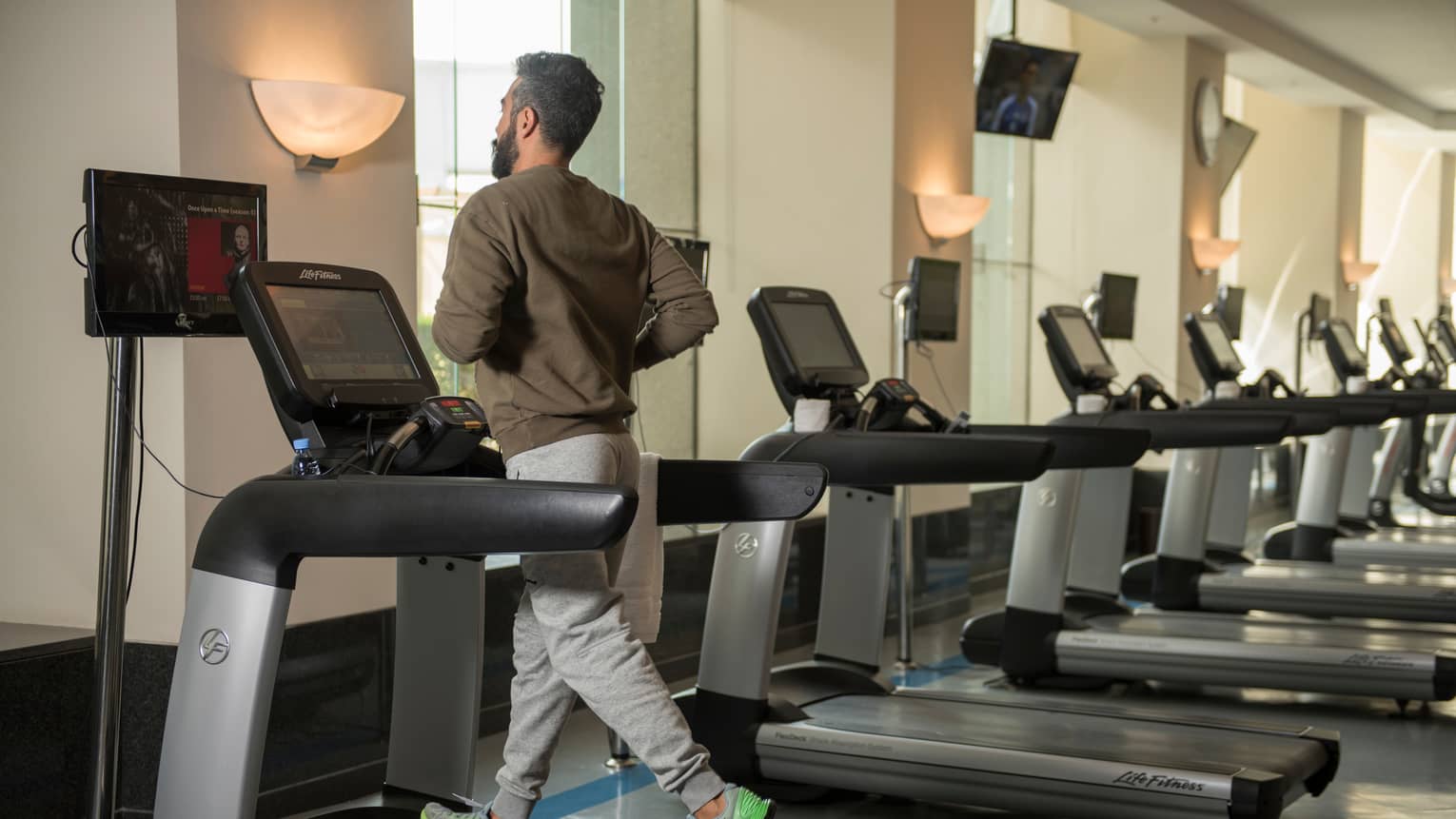 Back of man in sweatshirt and sweatpants as he runs on treadmill in gym next to row of cardio machines