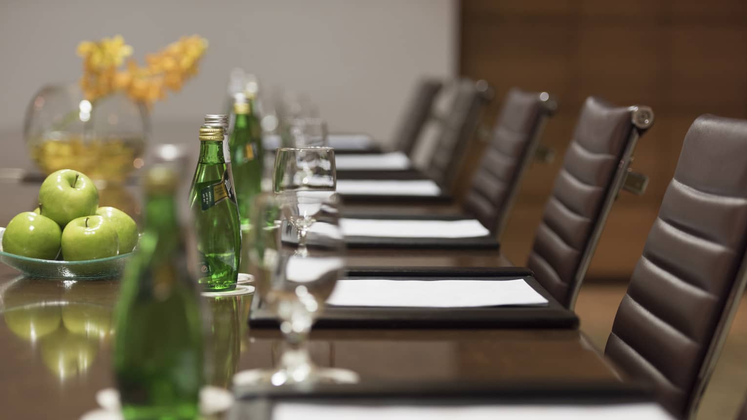Close-up of business meeting table row of chairs, paper agendas, green glass bottles, apples