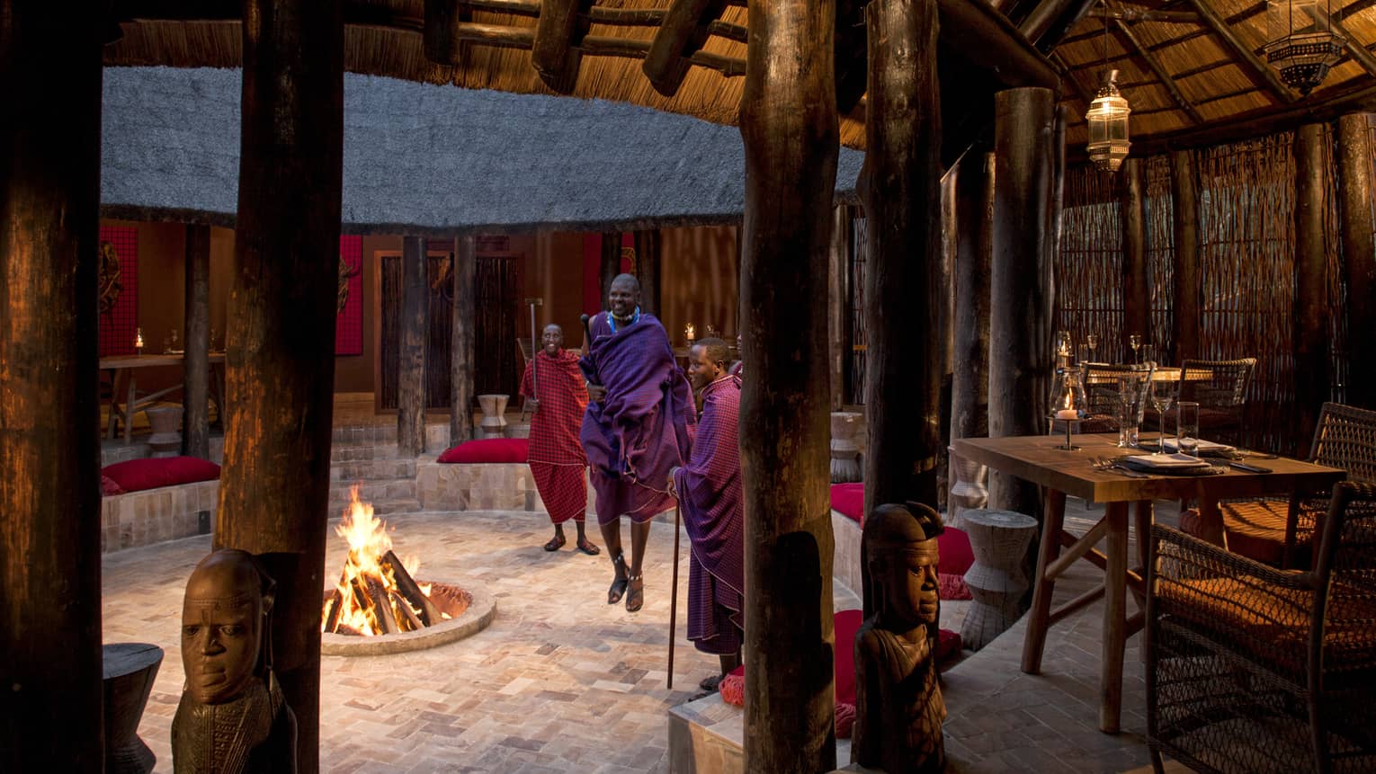 Maasai perform around a fireplace under a thatched hut with rustic tables and chairs at Boma Grill