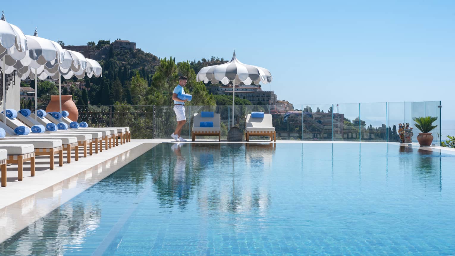 A person walks near the end of a long infinity-edge pool that has lounge chairs and umbrellas along one side and the end, with land and ocean views in the background