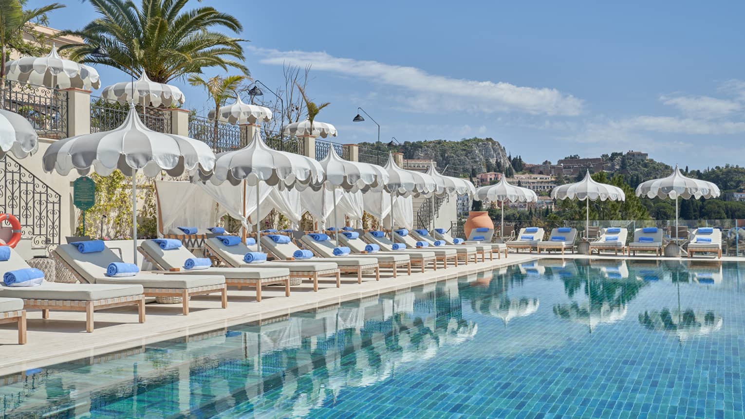 A long, smooth infinity-edge pool with lounge chairs and umbrellas along one side and one end. Palm trees and hilly landscape views are in the background.
