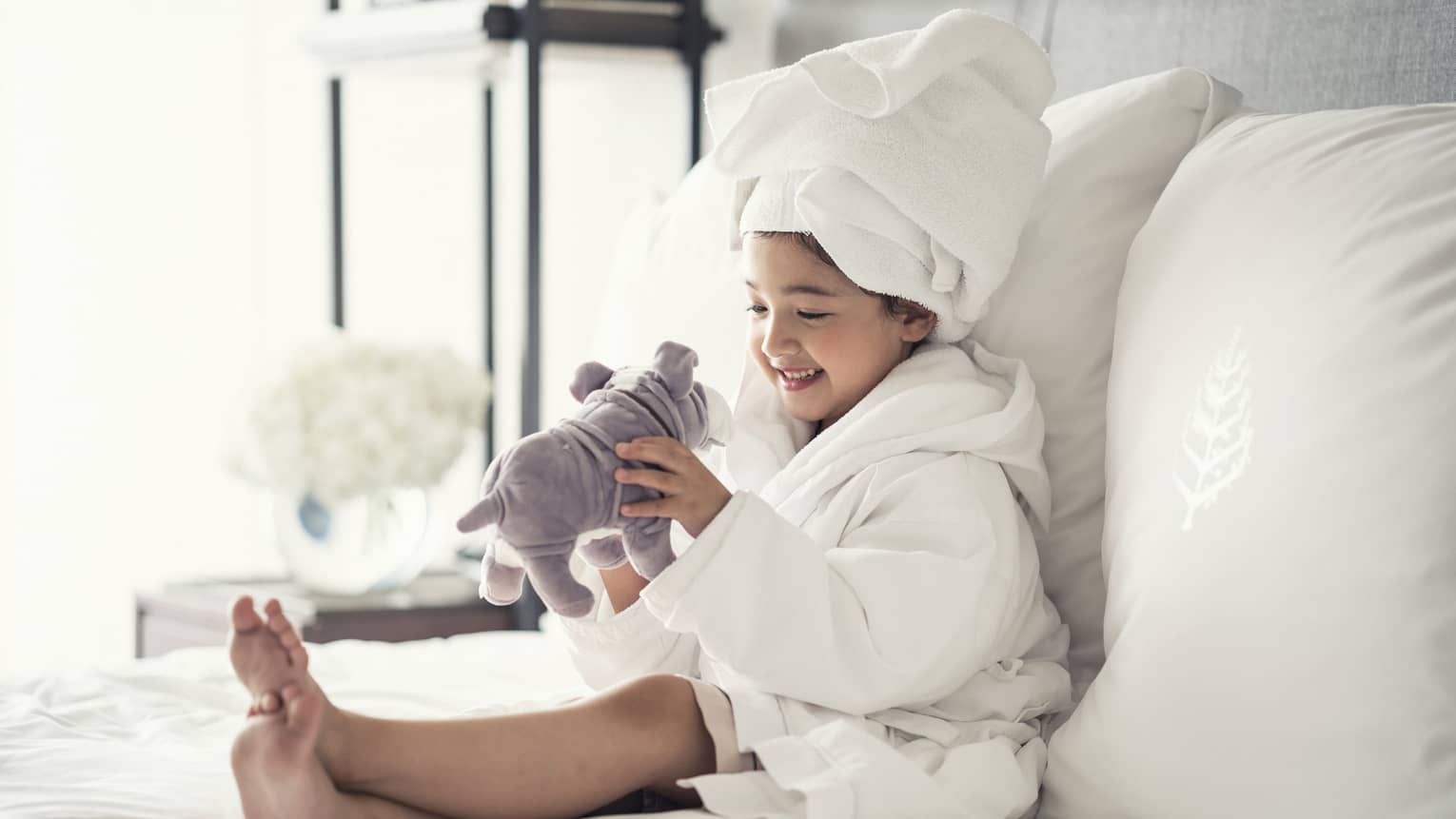 Young girl wearing a white bathrobe with towel wrap plays with plush dog toy on bed