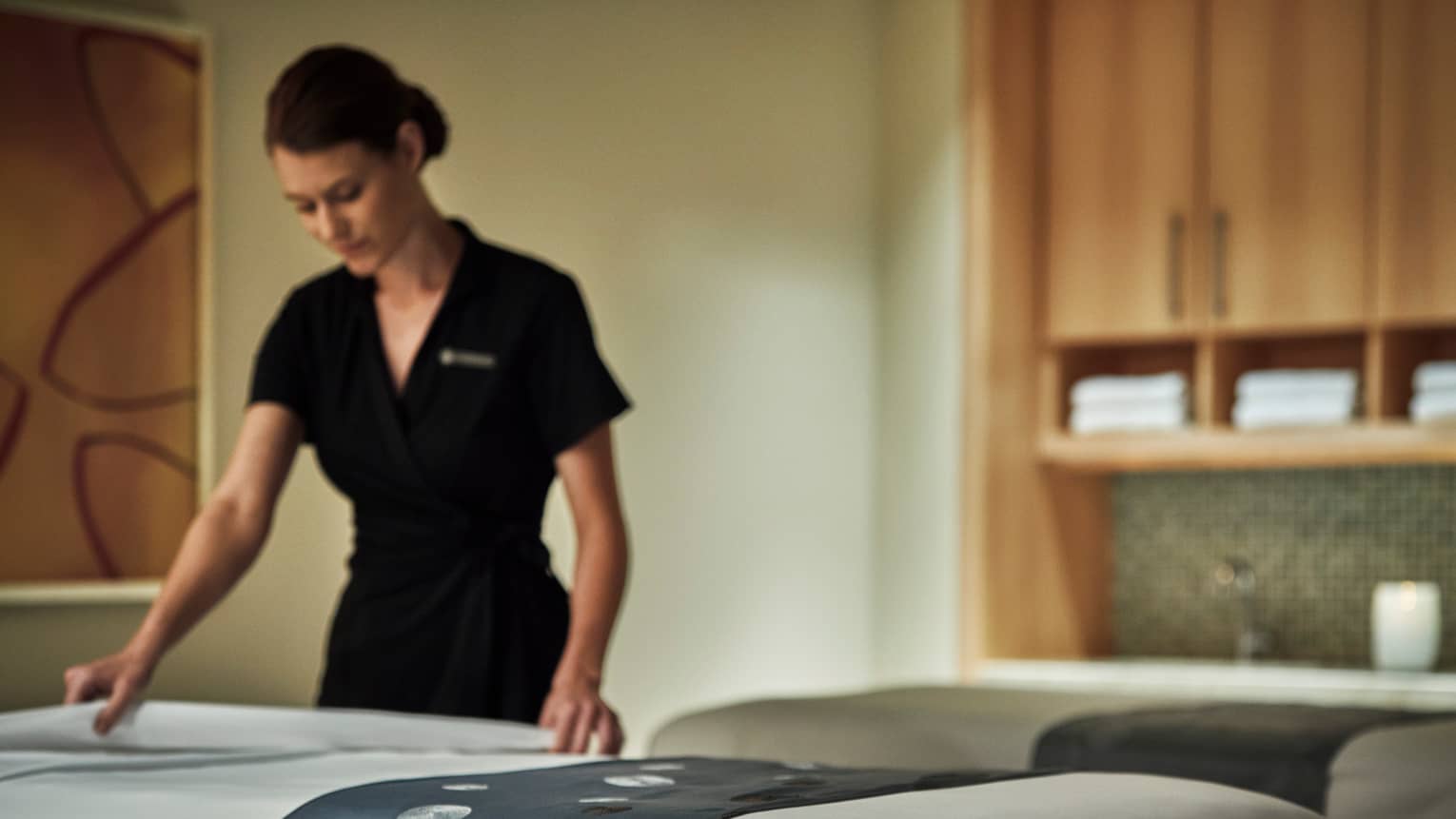 Spa attendant in black dress folds down white sheet on massage table