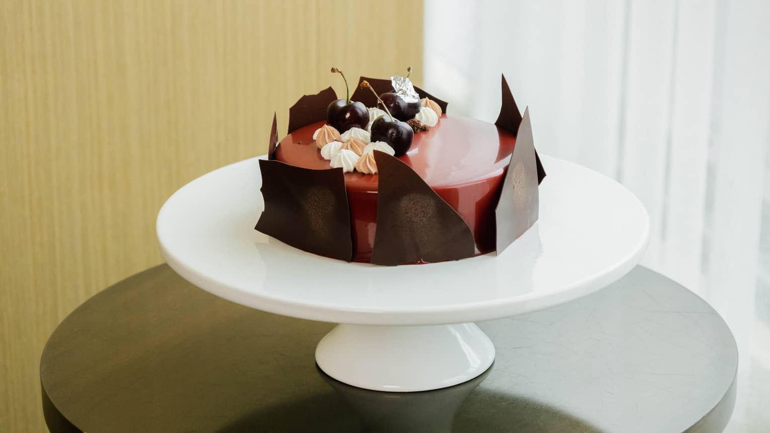 Round black forest cake with a red glazed center, jagged pieces of chocolate lining the sides, and fresh cherries and dollops of mousse is set atop a round white cake stand