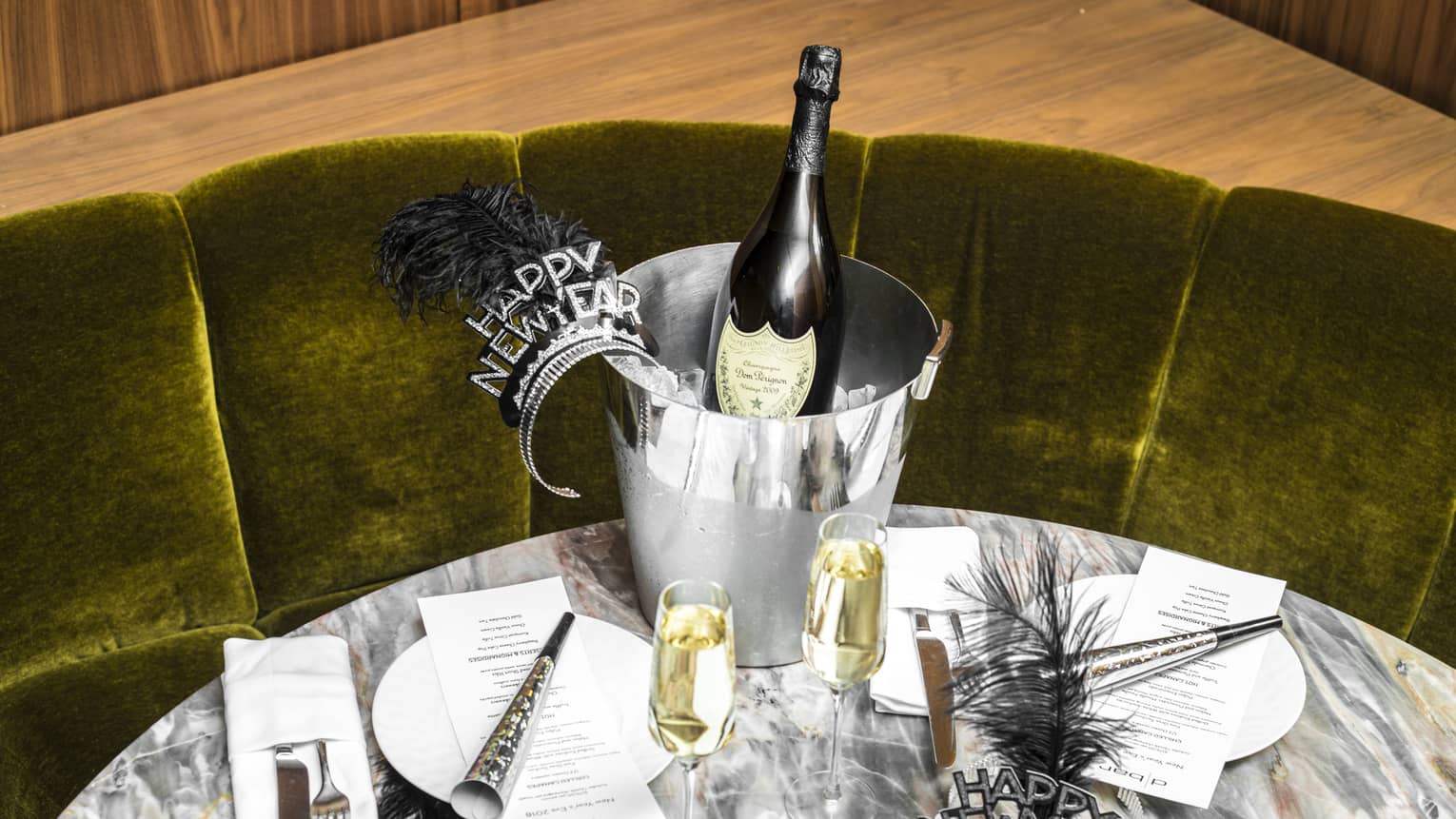 Green velvet banquette and a marble round table are set with New Year's Eve decorations, including two glasses of champagne, black feather headbands, cone-shaped noisemakers, a bottle of champagne sitting in a silver ice bucket and two sets of plates and cutlery