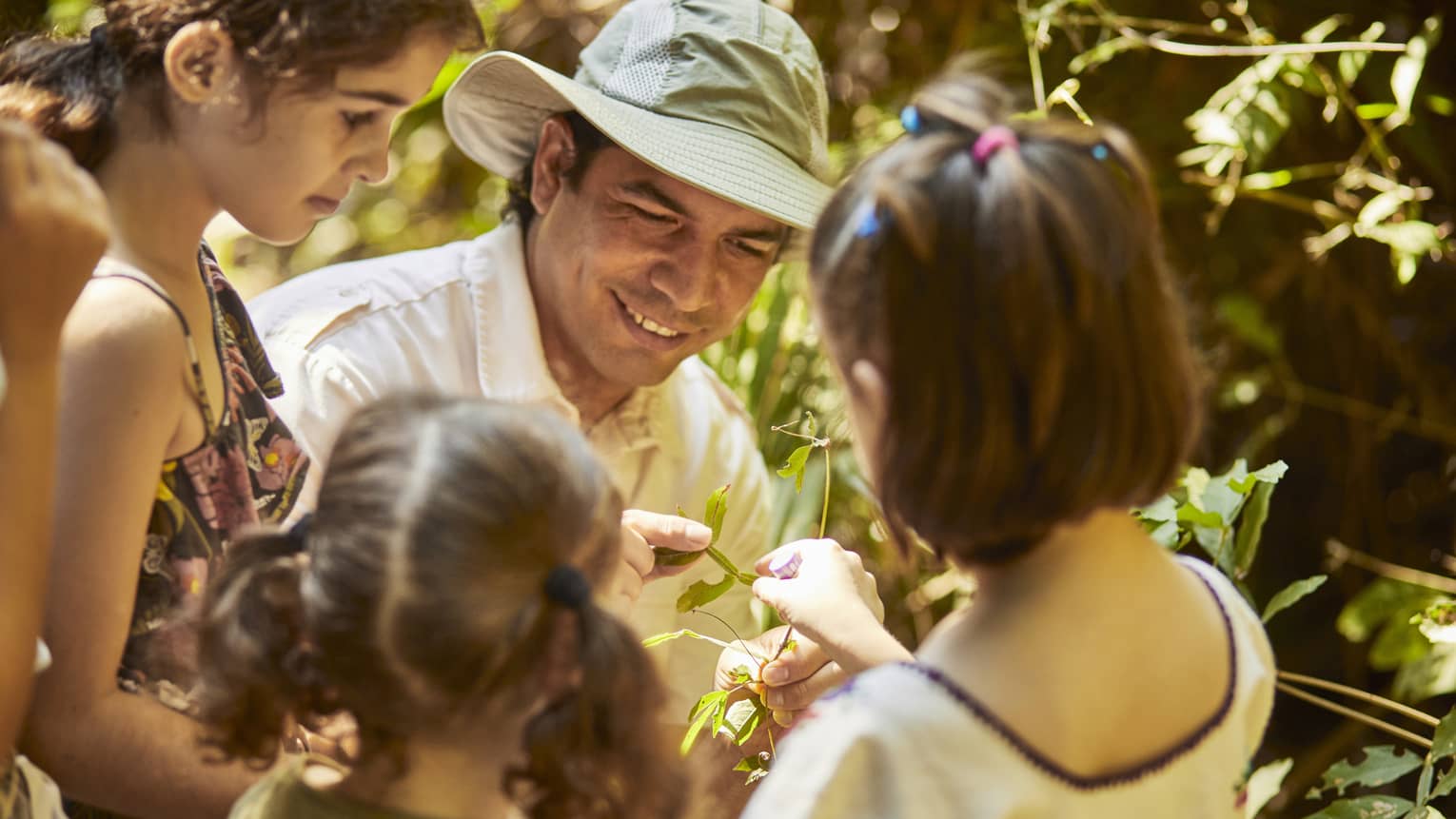 Three children gather around a guide wearing a sun hat as he shows them a plant. One child reaches to touch the leaves.