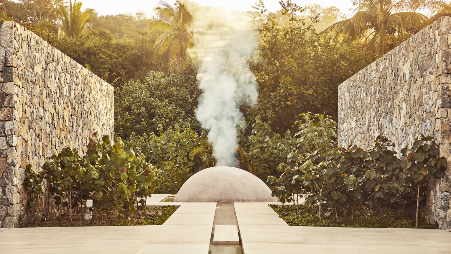 White steam rises from a small cement dome nestled between two large stone walls and surrounded by dense dark-green foliage.