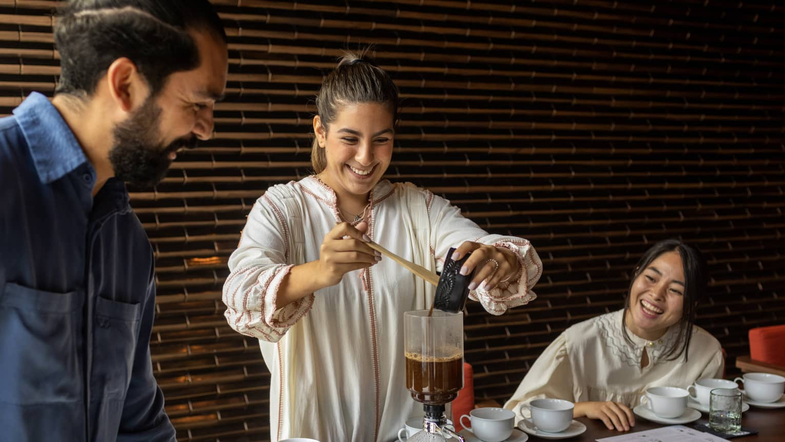 At a table set with white cups and saucers, two smiling guests watch as another pours coffee into a siphon coffee maker.
