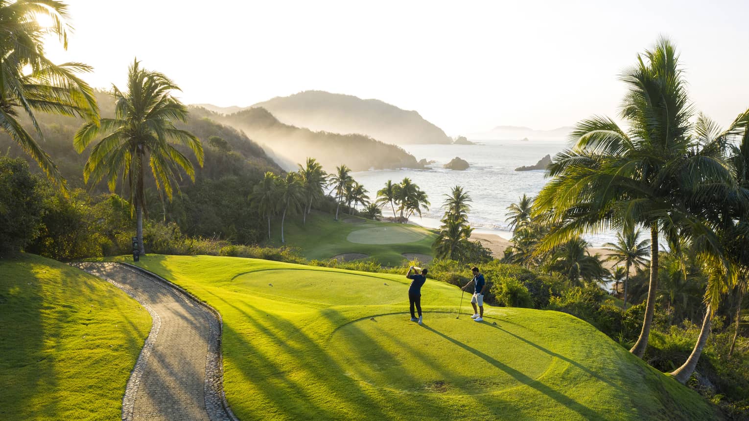 Rear view of two golfers on an elevated tee, one swinging toward the green down by the beach, misty hills and ocean beyond.
