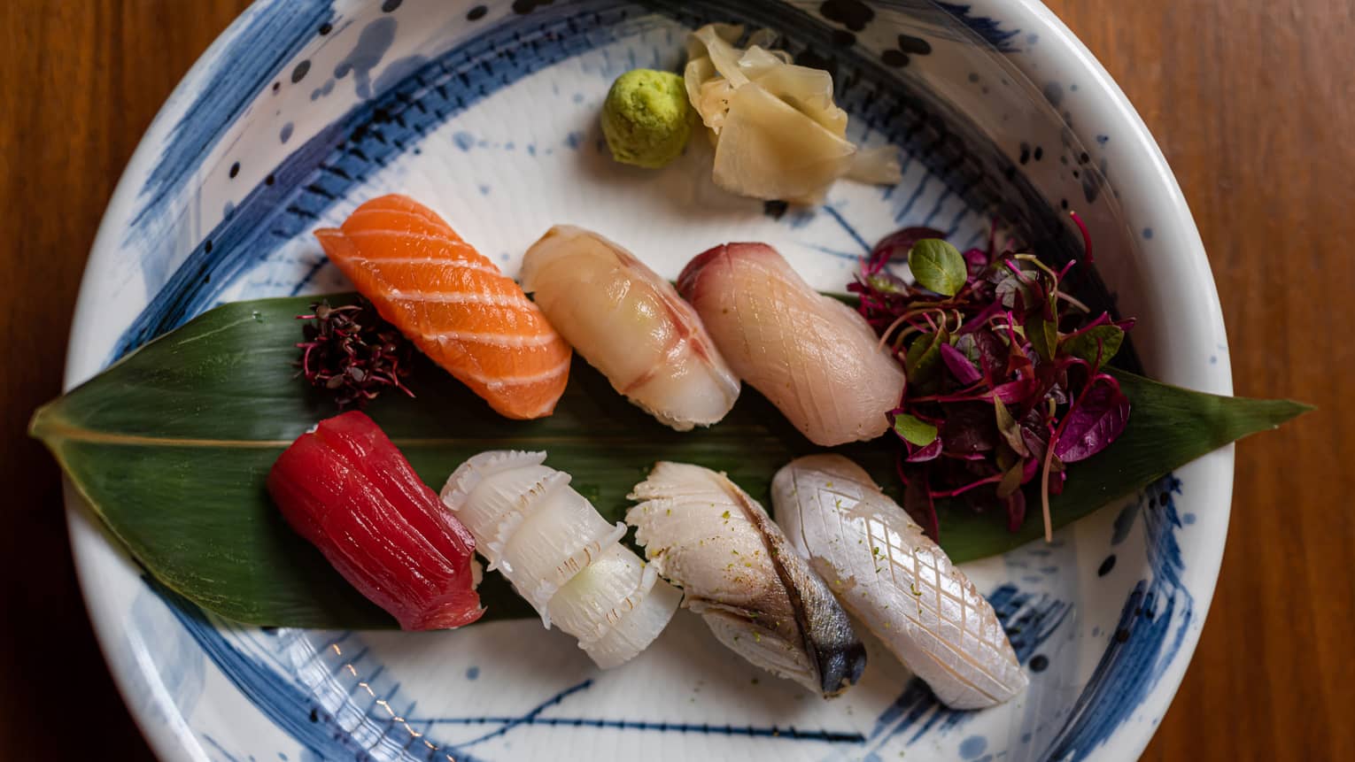 A round blue-and-white plate holds seven colourful pieces of nigiri sushi on a large leaf, wasabi and ginger on one side. 
