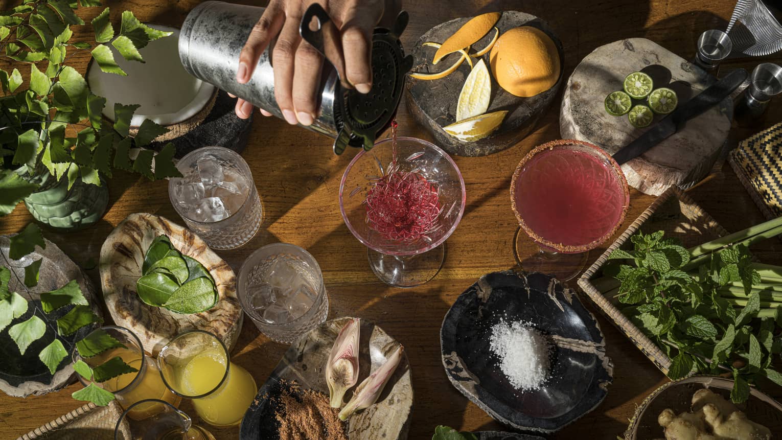 Aerial view of bartender's hand pouring ruby-coloured cocktail intro stemmed glass, local ingredients on wooden bartop surrounding it