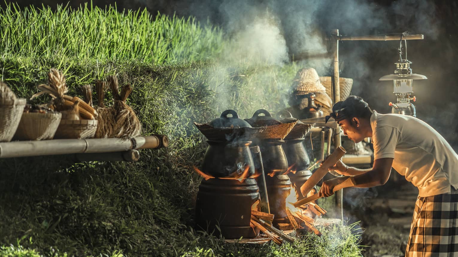 A chef stokes the fire beneath a row of outdoor cooking pots set on a grassy hill, the smoke illuminated by the flames.