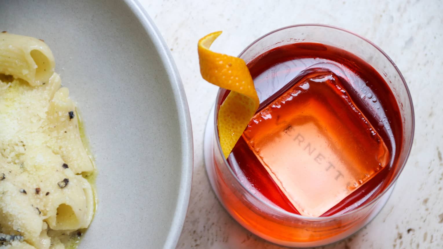 Glass with a red coloured drink beside a plate of pasta.