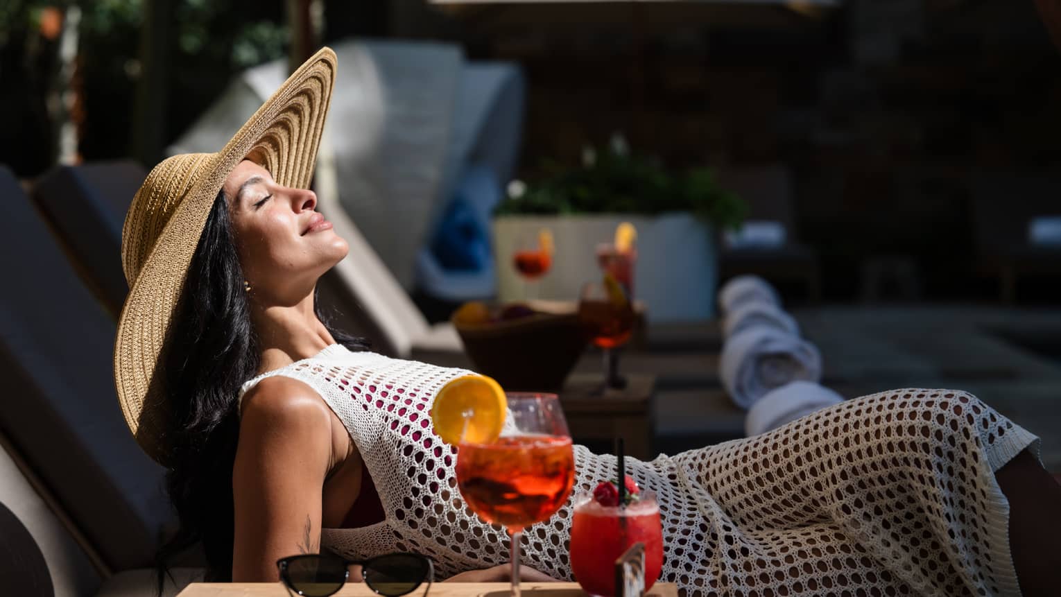 A guest laying in a lounge chair in the sunshine with their sunglasses and drink on a table beside them