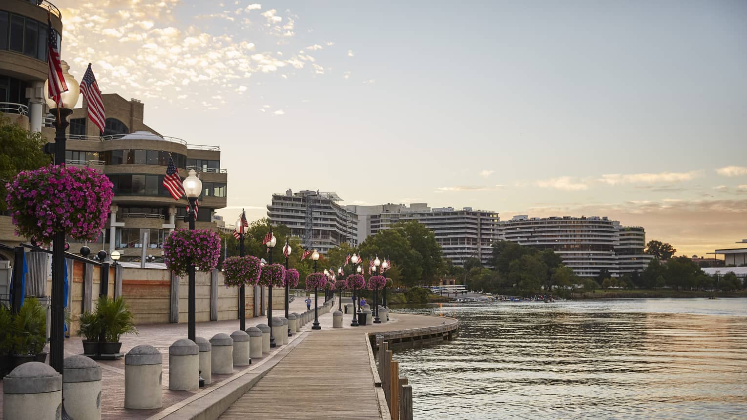 A waterfront with a wooden walkway next to light posts and flowers with building on the other side of a small road.