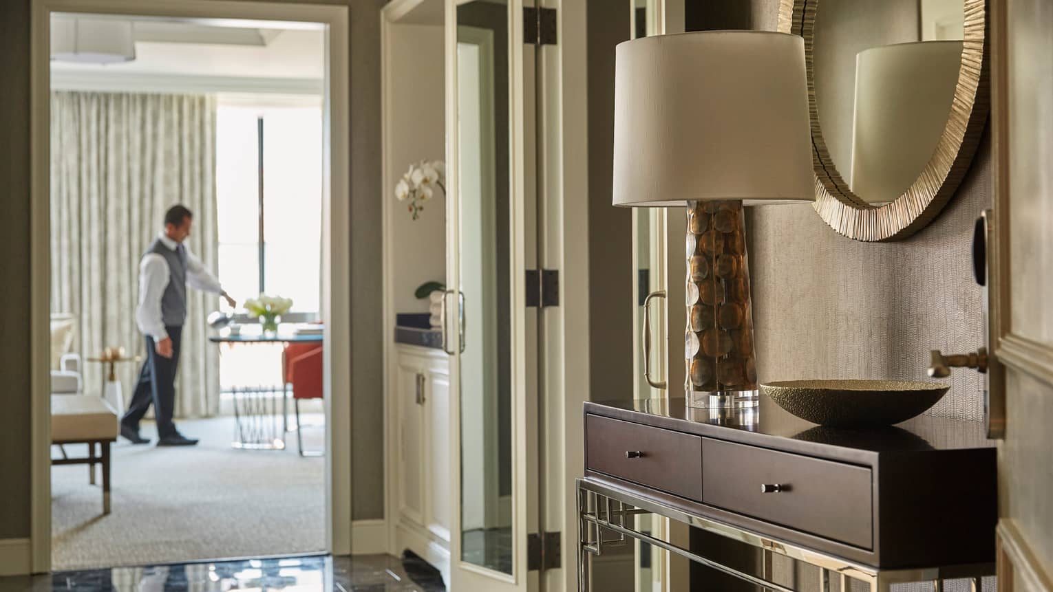 View from hotel suite hall with console table, lamp past doors to staff setting in-room dining table