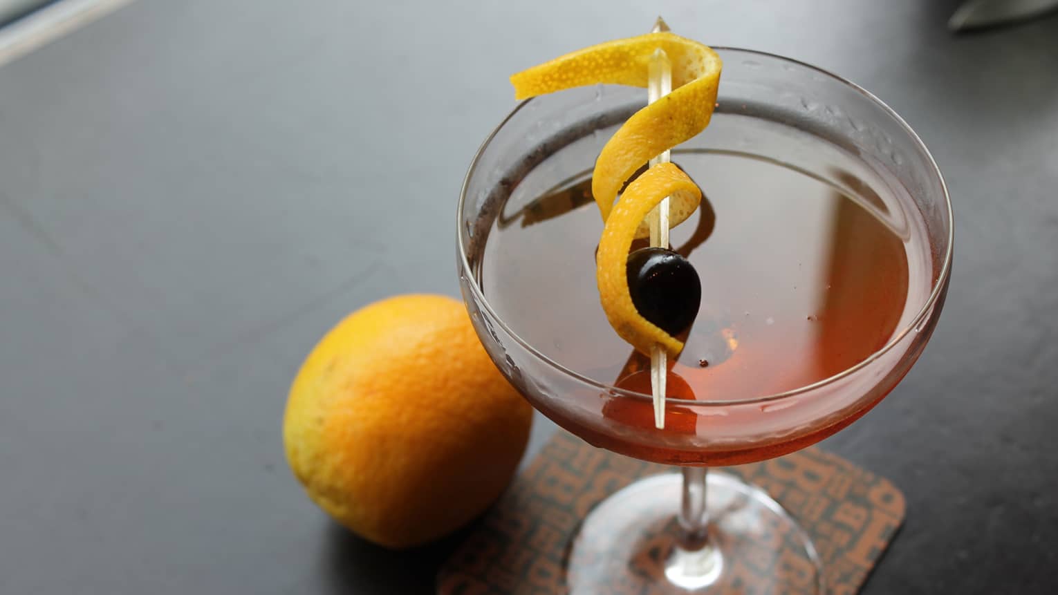 Bourbon cocktail in glass with cherry and orange rind garnish on toothpick, whole orange on bar