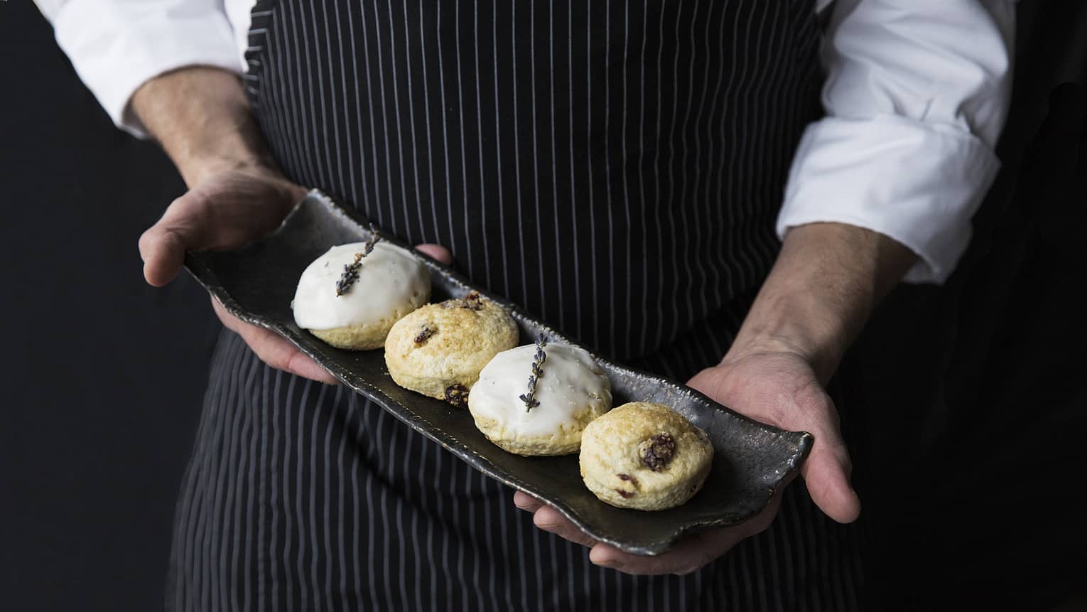 Chef wearing black striped apron holds rustic platter with four gourmet scones at Afternoon Tea 