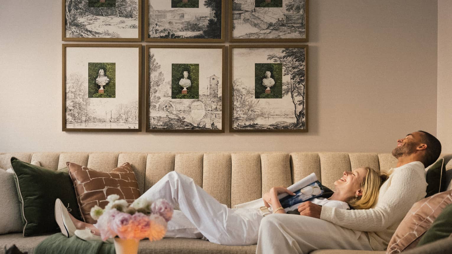 Two guests laying on the couch in a guest room with picture frames on the wall and a bouquet of flowers on the coffee table