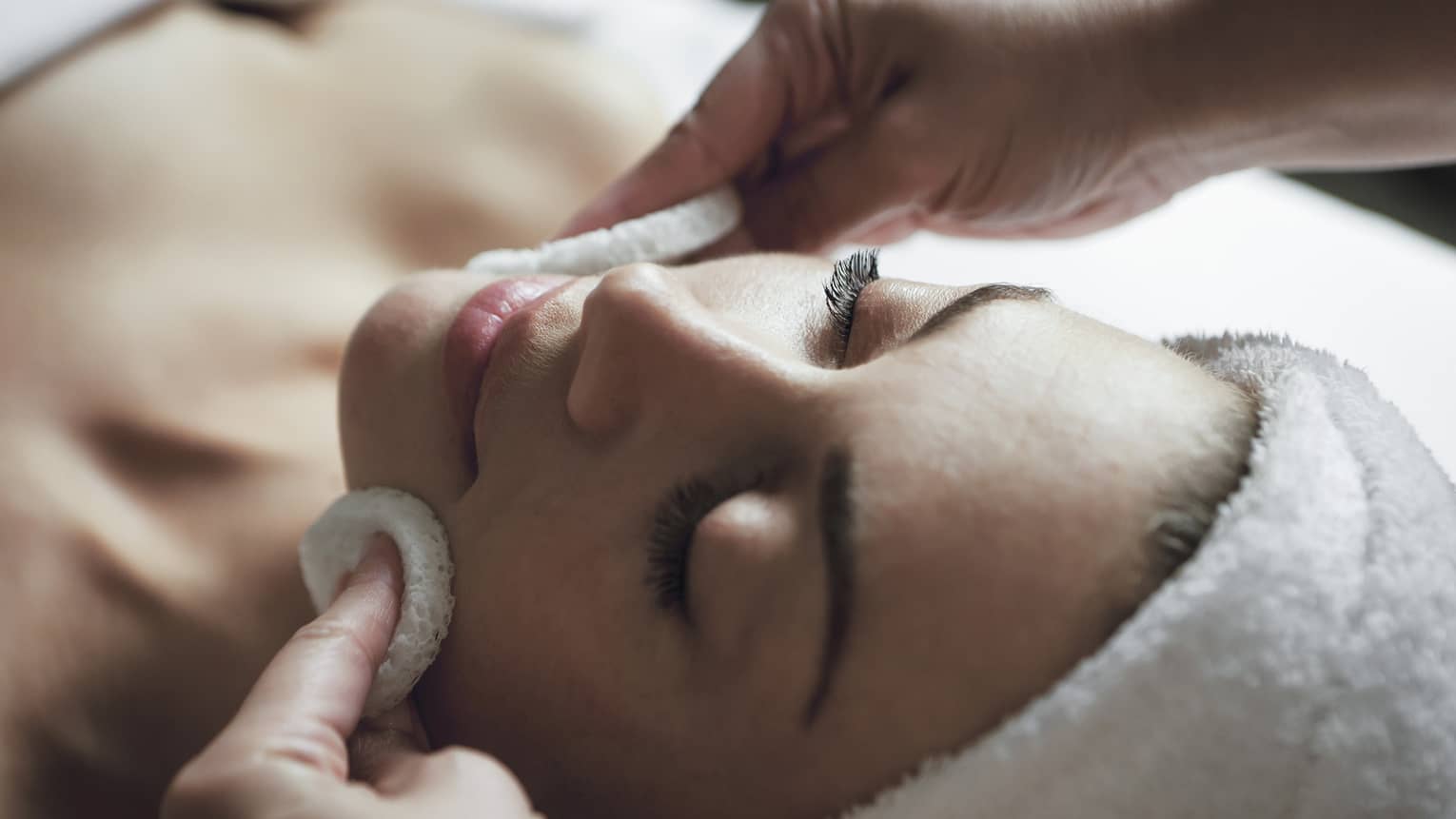 A detail of hands rubbing a sponge on a woman's face as she lies on a massage table with her eyes closed and hair wrapped up in a white towel
