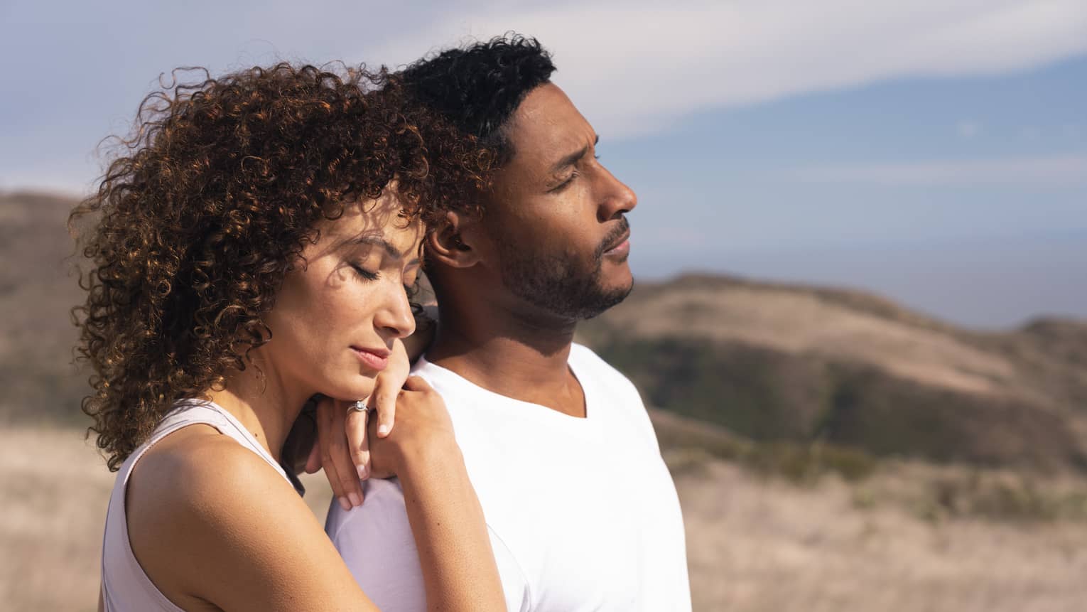 Two people both wearing white t-shirts stand together in a field with their eyes closed, one with their arms on the other's shoulders