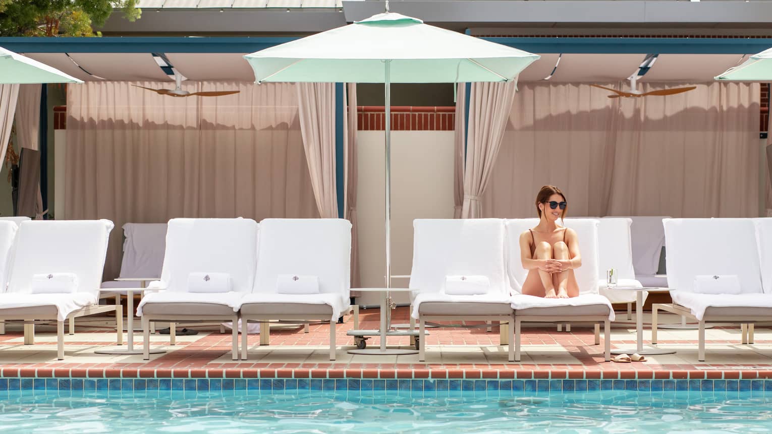 A woman resting on a lounge chair under an umbrella near an outdoor pool.