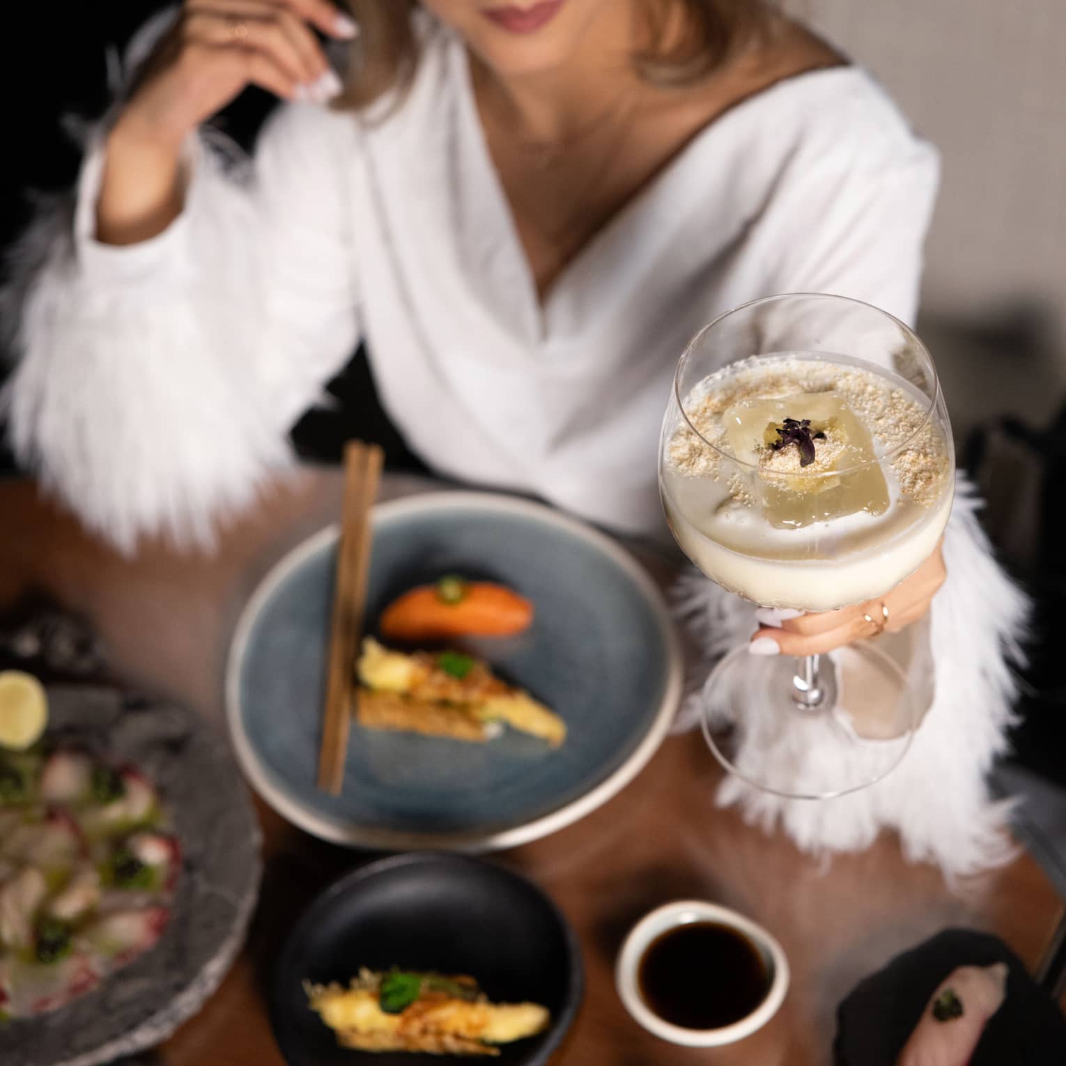 Person wearing white dress with feathered sleeves holds up a white cocktail in a large stemmed glass