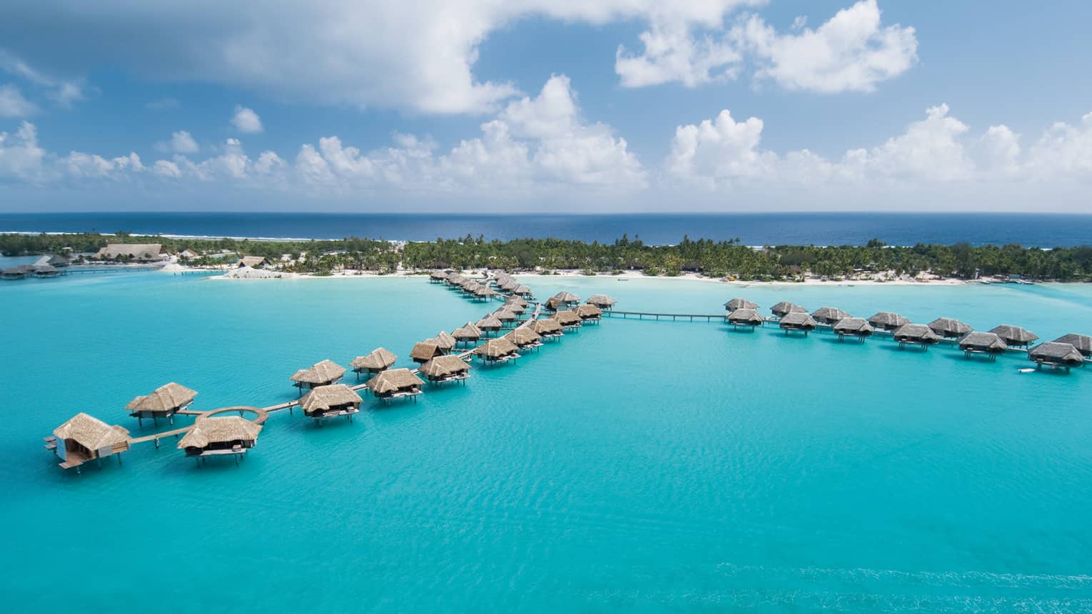 Aerial view of two rows of overwater bungalows far out in lagoon with resort island in background