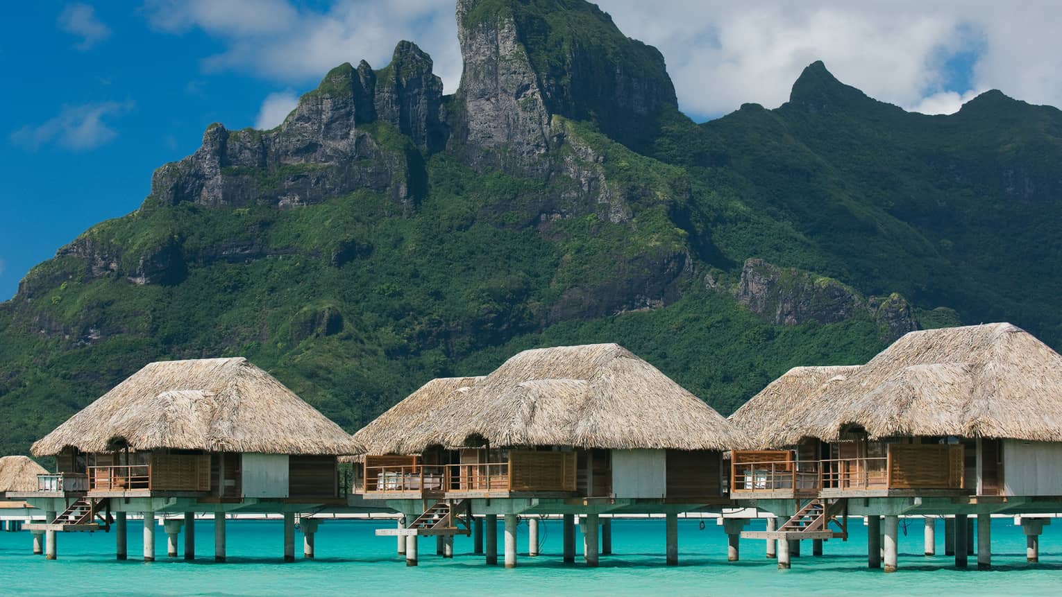 Close-up exterior view of six overwater bungalows over blue lagoon, mountain in background