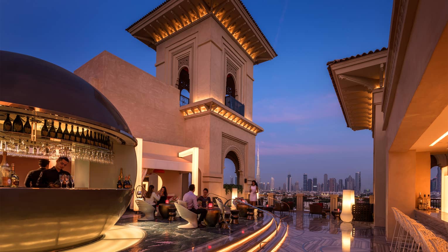 Rooftop lounge at night, with modern gold bar and guests sitting on white chairs, city skyline visible