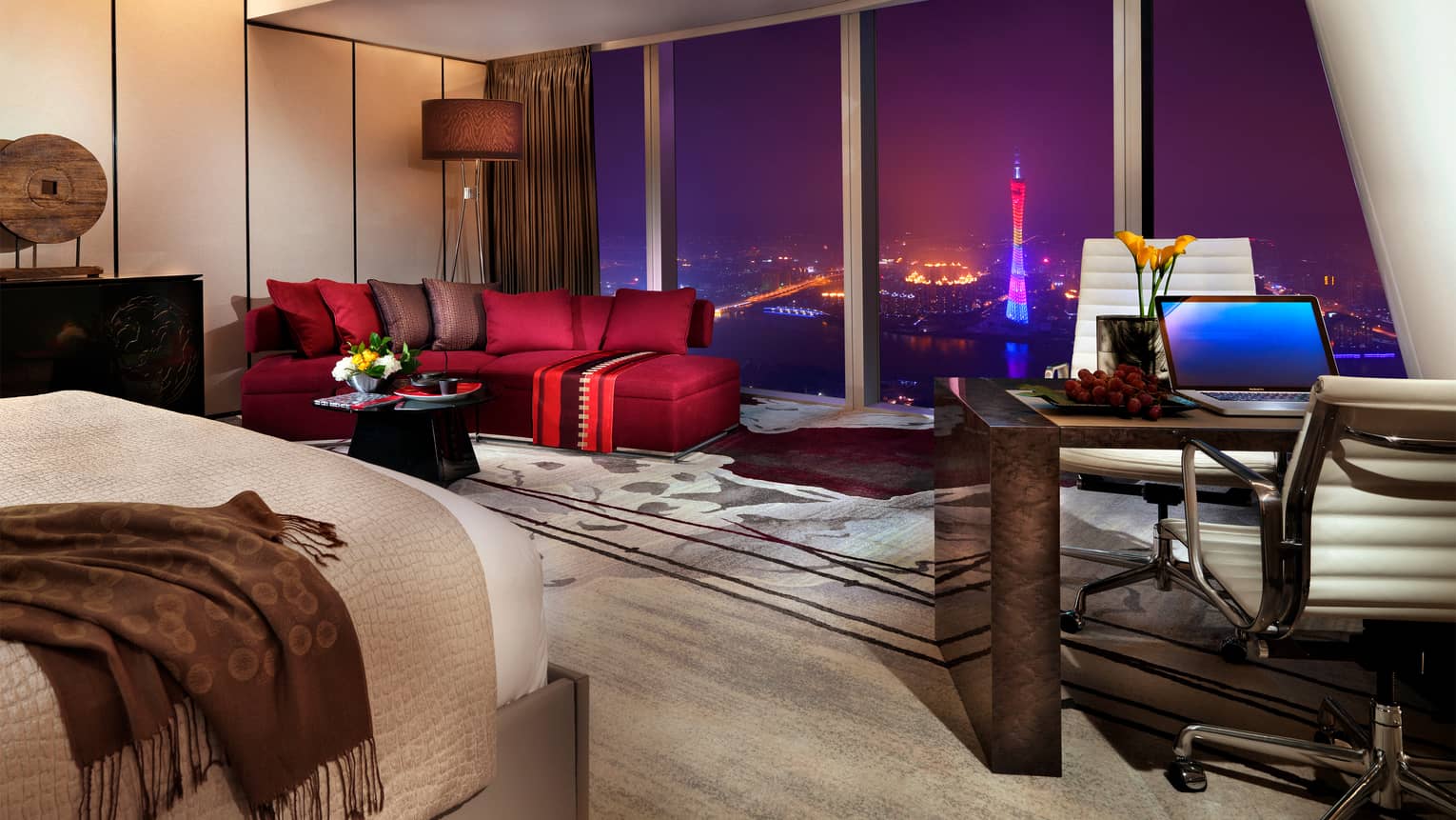 Floor-to-ceiling hotel room window at night with city view, beside stylish red sofa and area rug