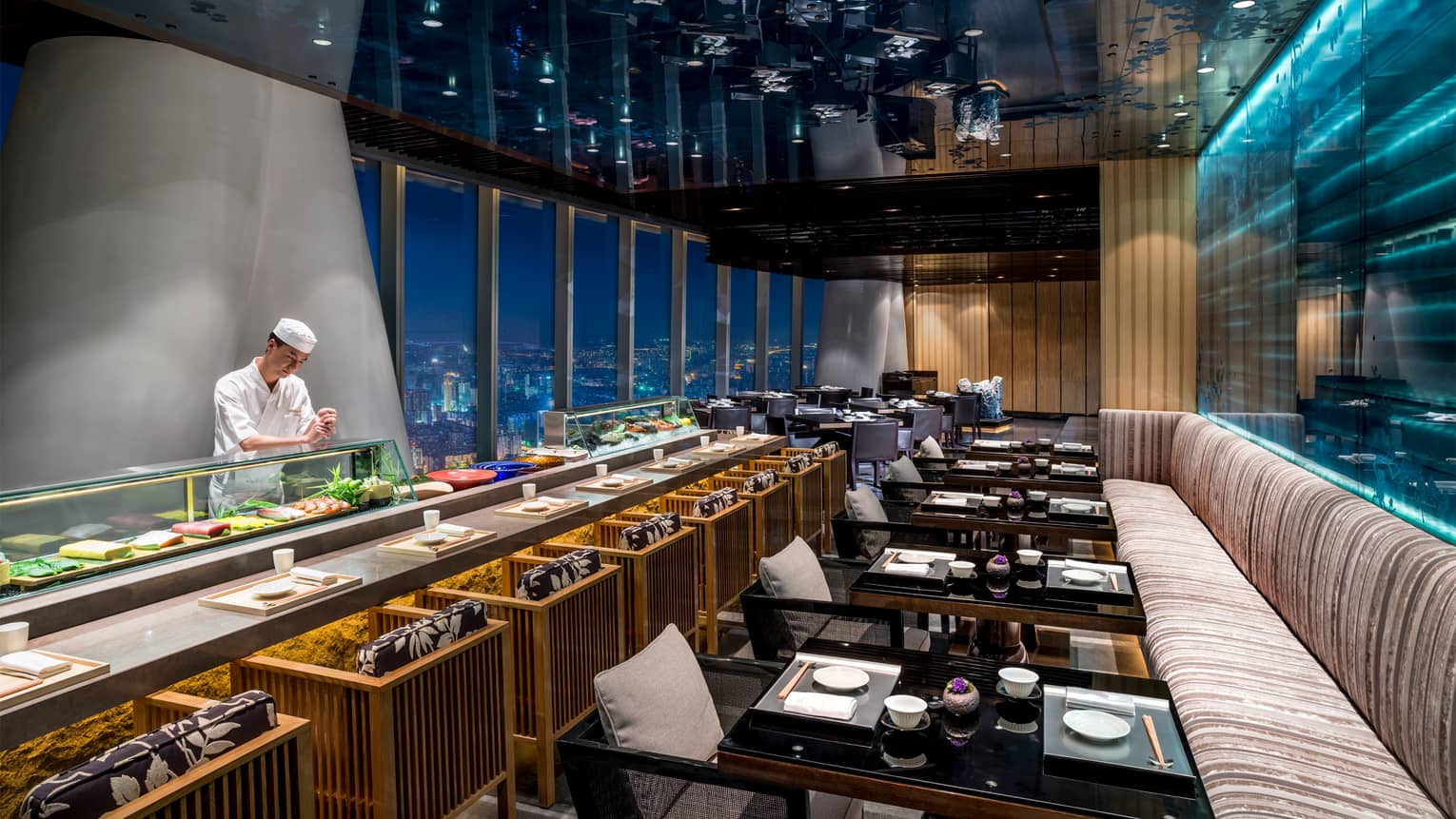 Kumoi Japanese restaurant chef stands behind long sushi bar in dining room overlooking city lights