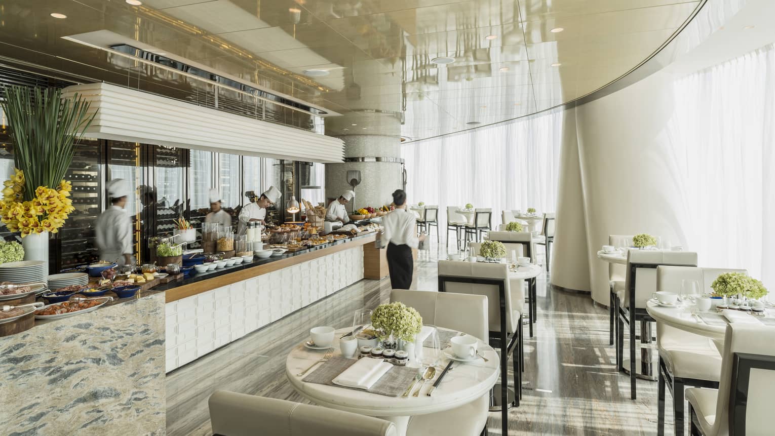 Restaurant dining room with an open live-action kitchen, white walls and chairs, natural light