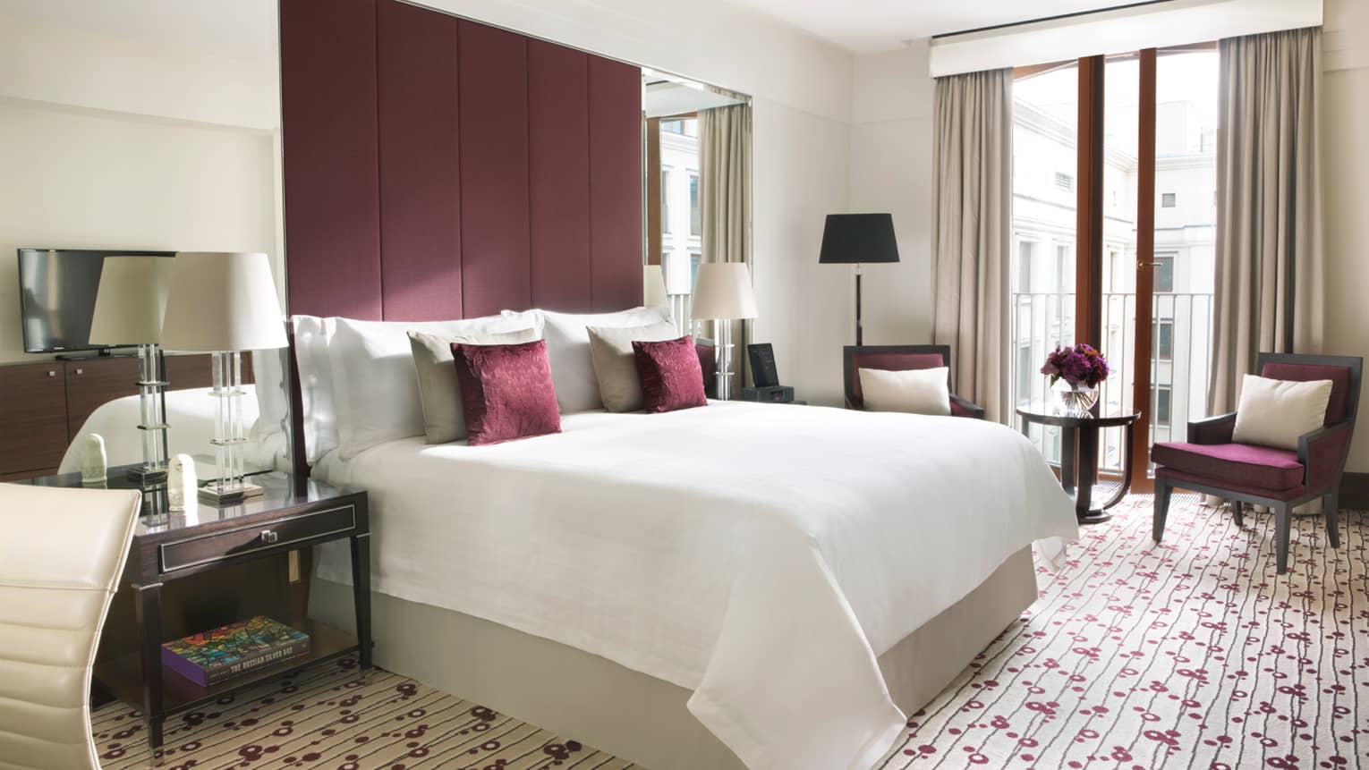 Deluxe Room bed with burgundy fabric headboard, accent pillows and chair, purple flowers in glass vase