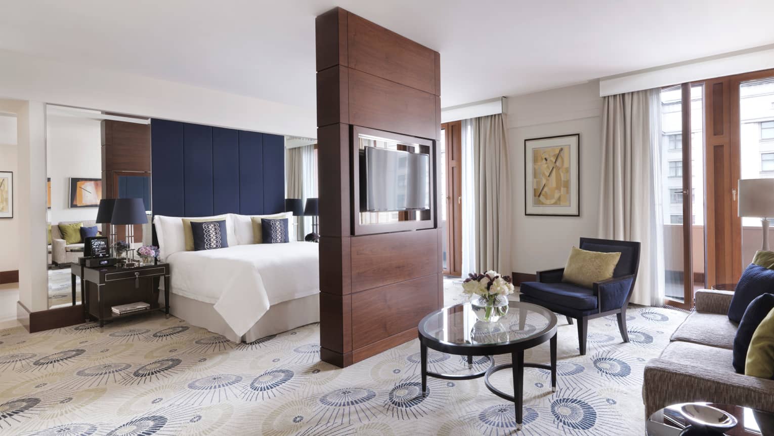 Suite with glass coffee table, wood room divider with TV, bed with blue fabric headboard and tall mirrors