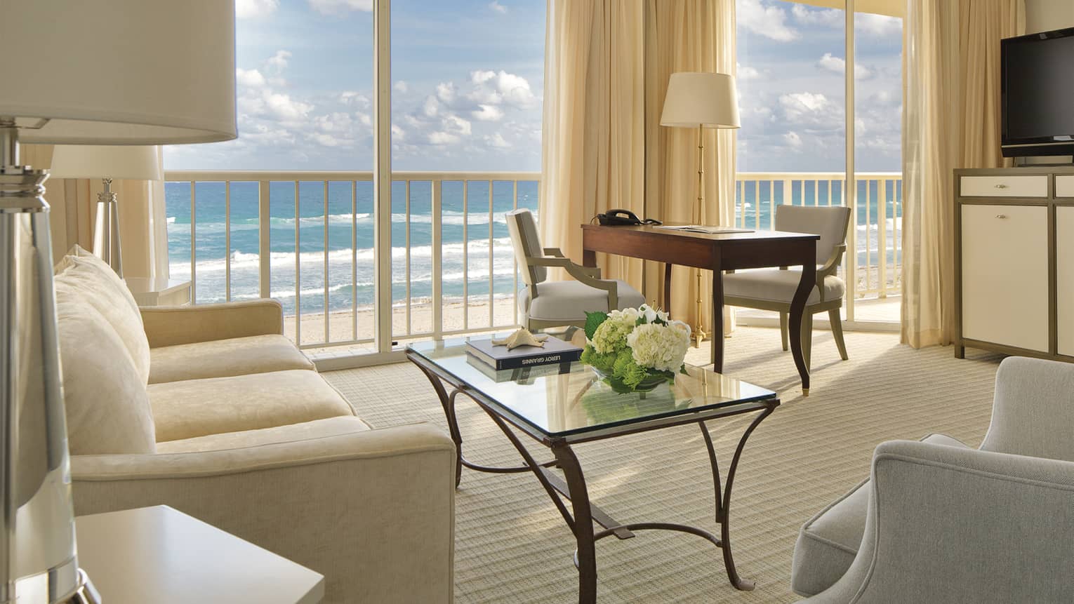Oceanfront Suite with white sofas and chairs, office desk with lamps, floor-to-ceiling balcony windows