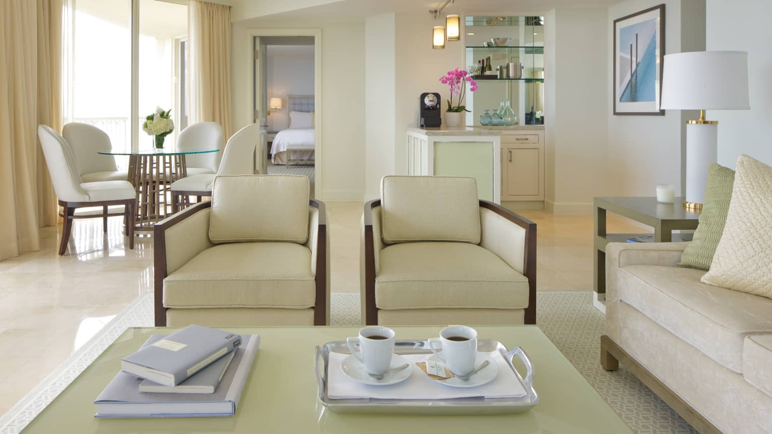Two white coffee mugs and saucers on tray beside books on coffee tables, white and cream-coloured chairs