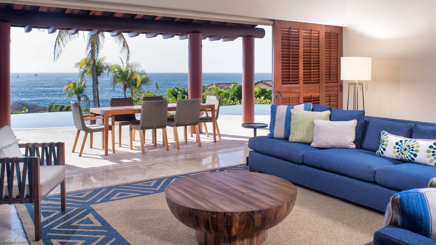 Large indoor-outdoor living room with blue sofa, round wood cofee table, open wall to outdoor dining table