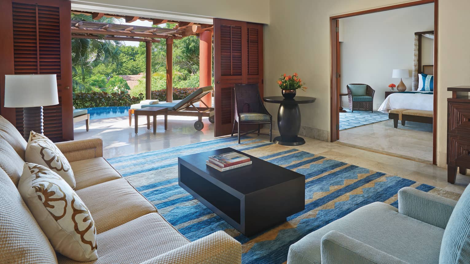 Garden view suite living area with full-size sofa and armchair, looking into bedroom, open door to patio with pool