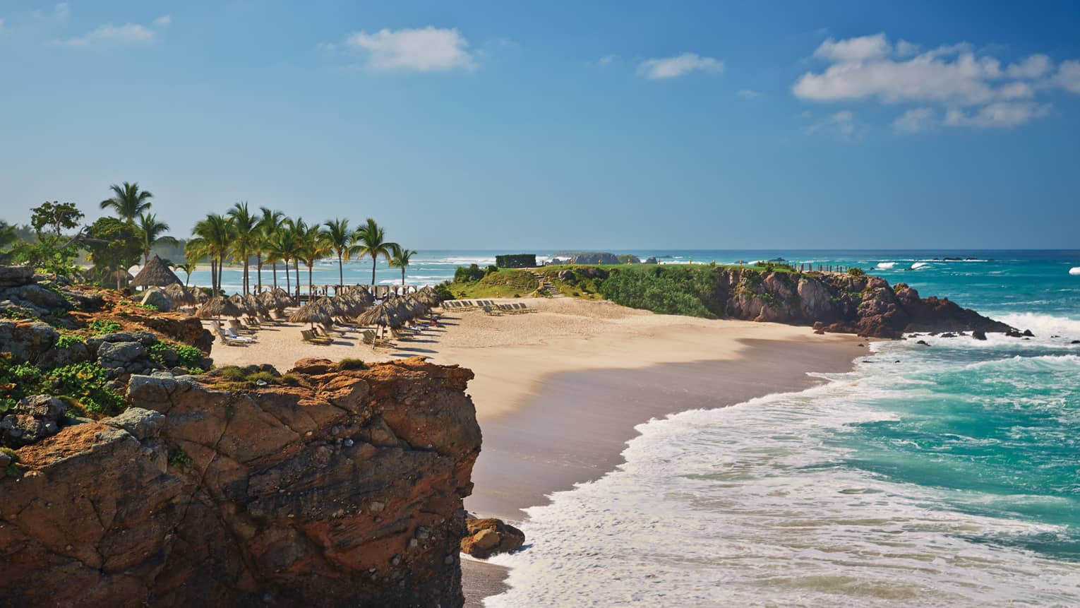 White sand beach lined with lounge chairs, palms, turquoise tides from oceanside cliff