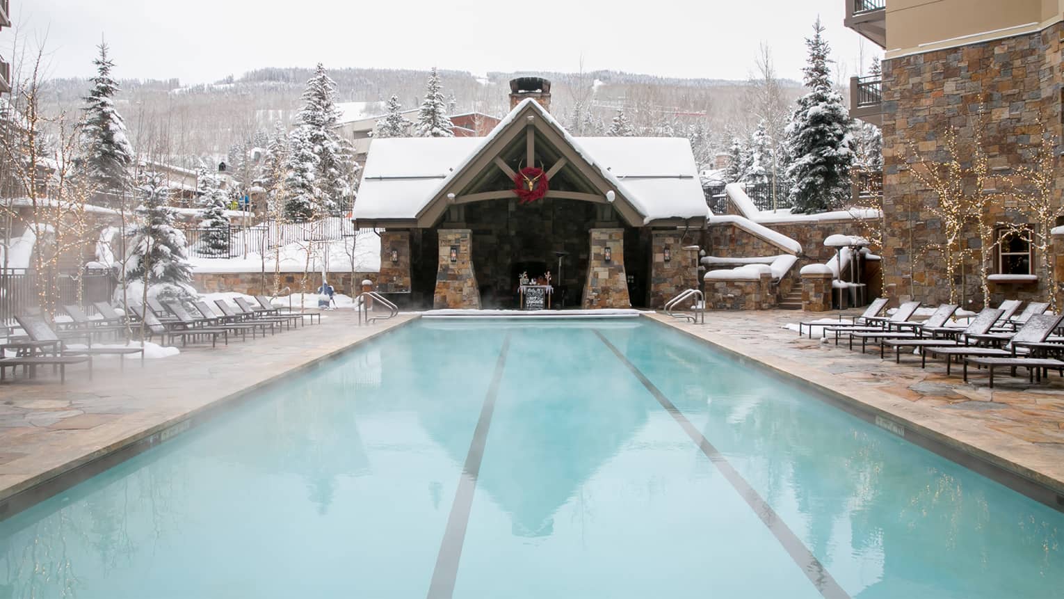 Steaming outdoor pool in winter with snow-covered lounge chairs, chalet-style cabana