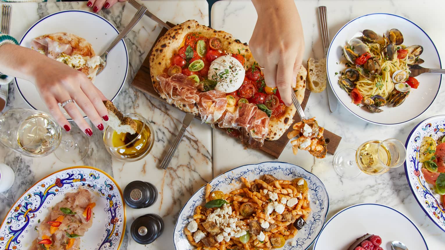A marble table features white wine and an array of colourful Mediterranean delicacies, hands reaching in to sample the food.