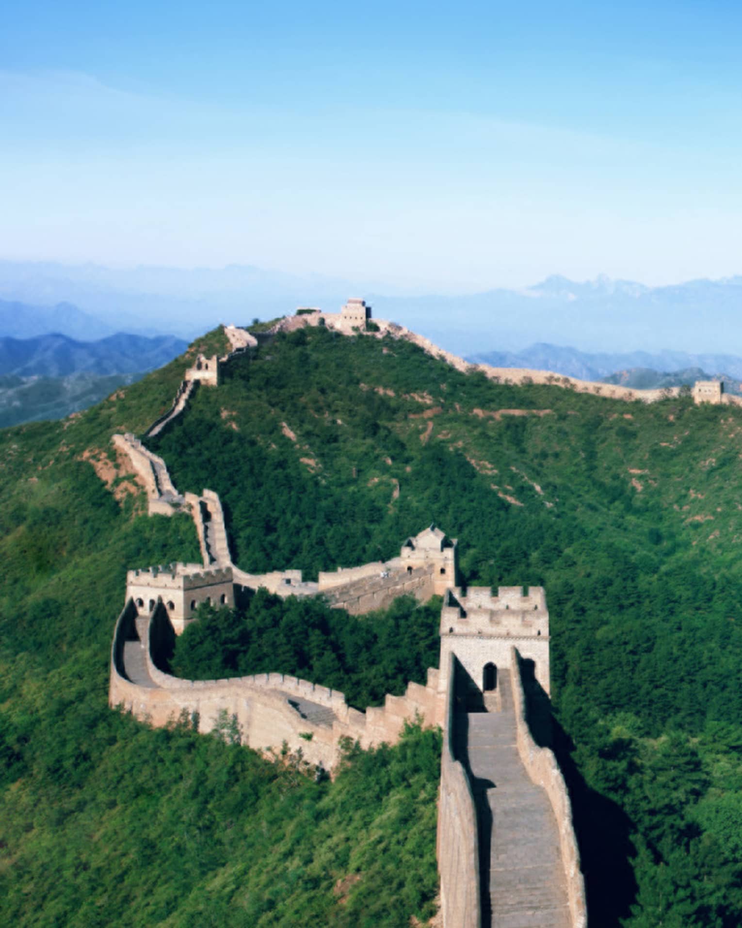 Stone watchtowers connected by stone walkways sit on the crestline of a lush, forested mountain range under a blue sky.