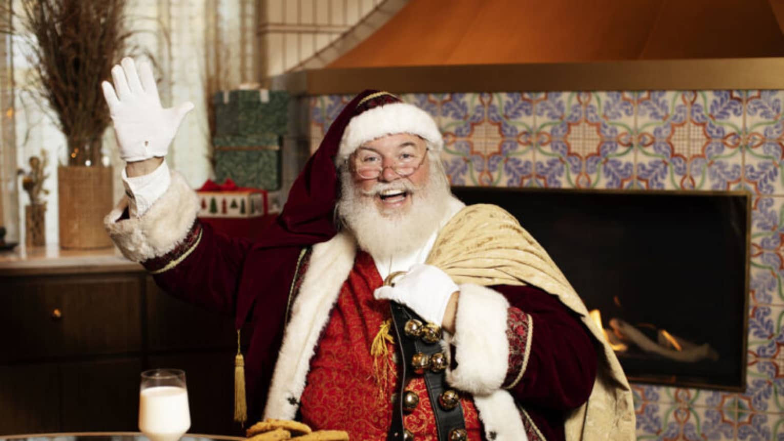 Santa Claus sitting at a table with a plate of cookies and glass of milk, and a fireplace in the background.