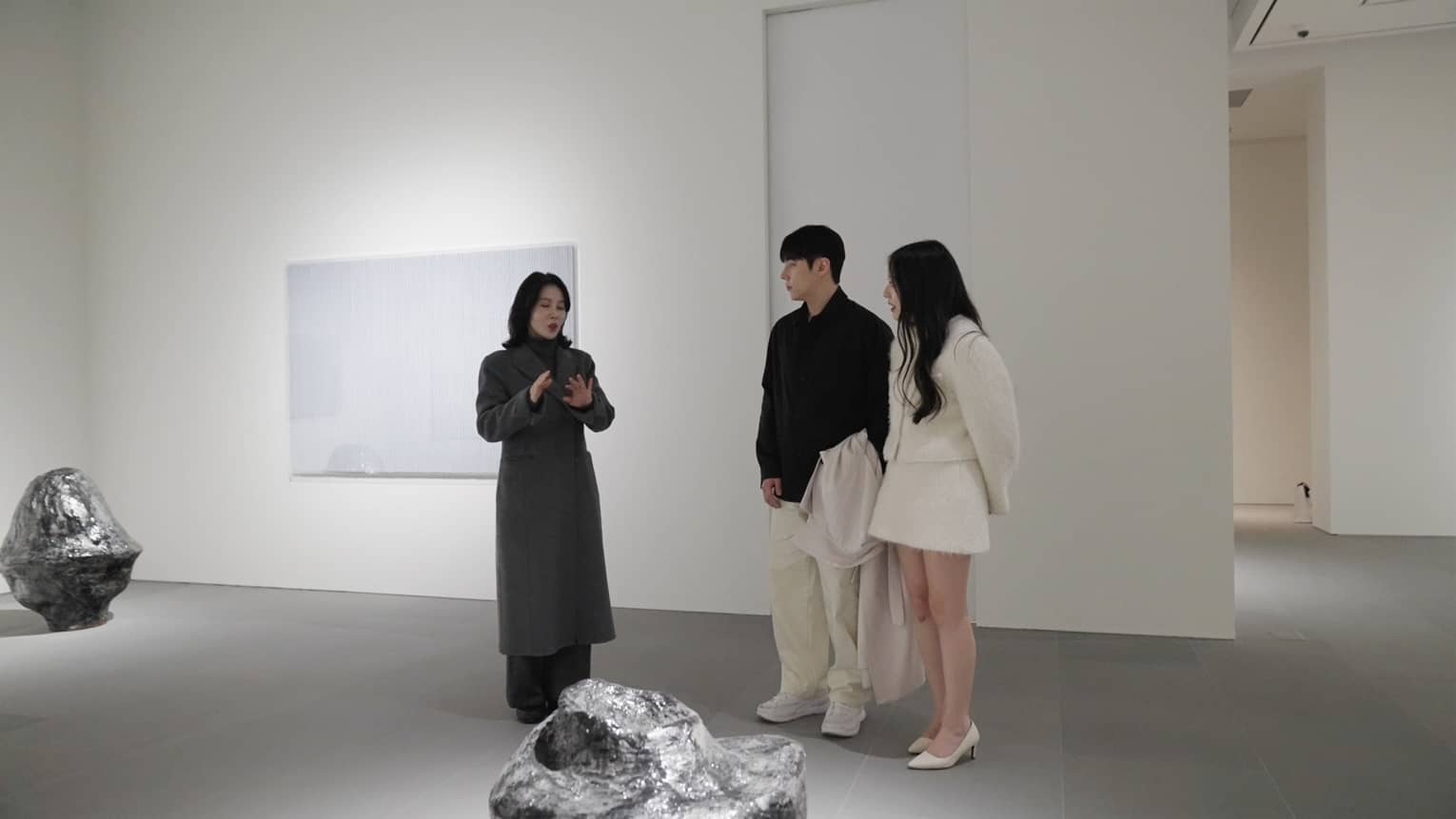 A couple standing in a modern art gallery with white walls and gray flooring watches as a person speaks about a modern art piece in front of them