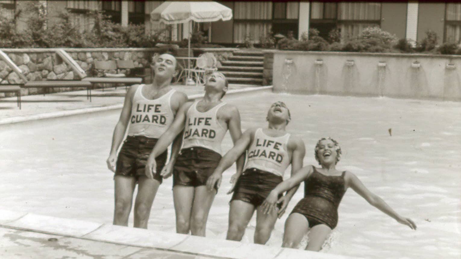 Black-and-white historic photo of three laughing lifeguards, smiling woman falling back into outdoor swimming pool